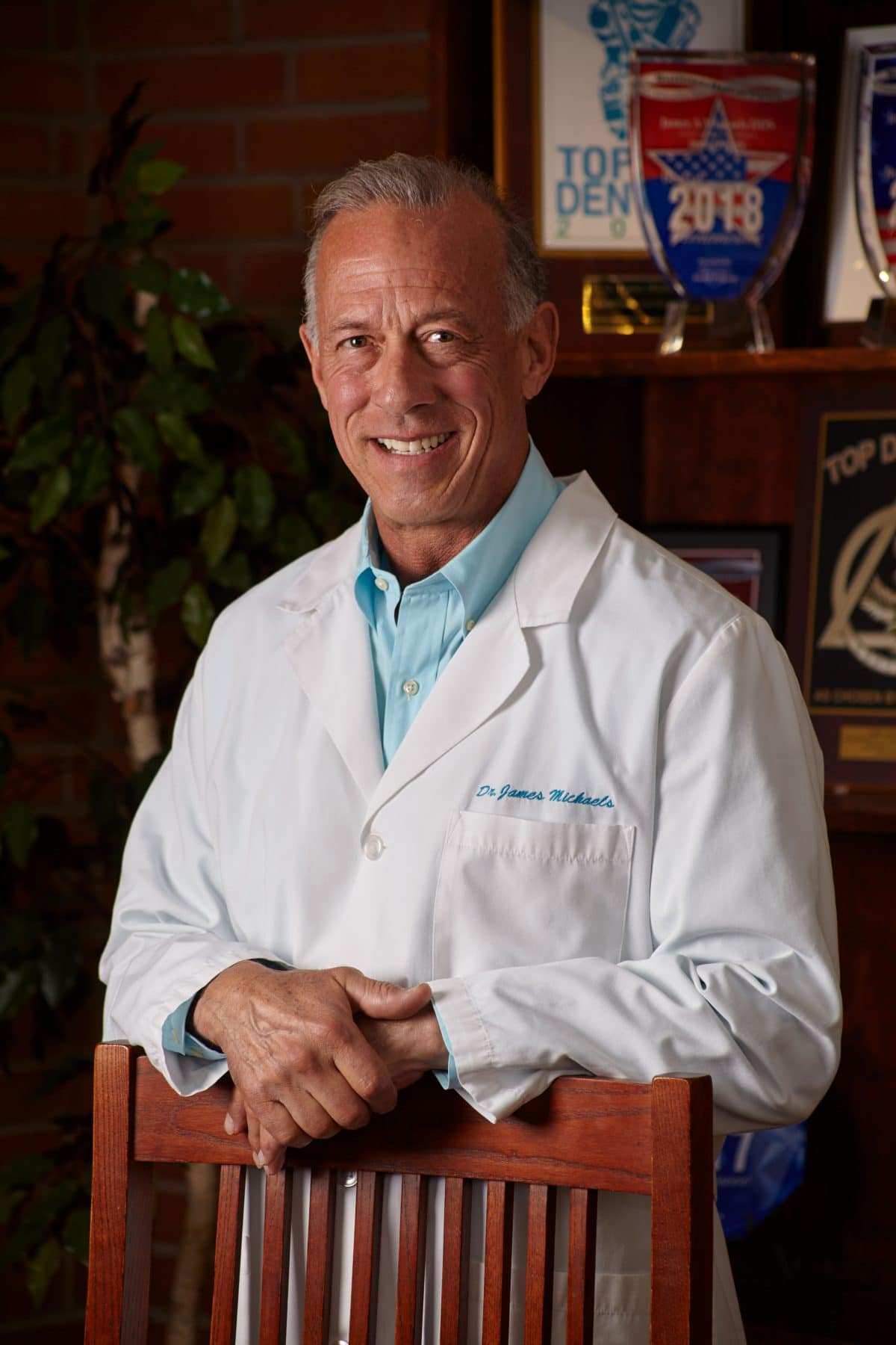 Man in white lab coat smiles, leaning on a wooden chair. Awards on shelf behind him.
