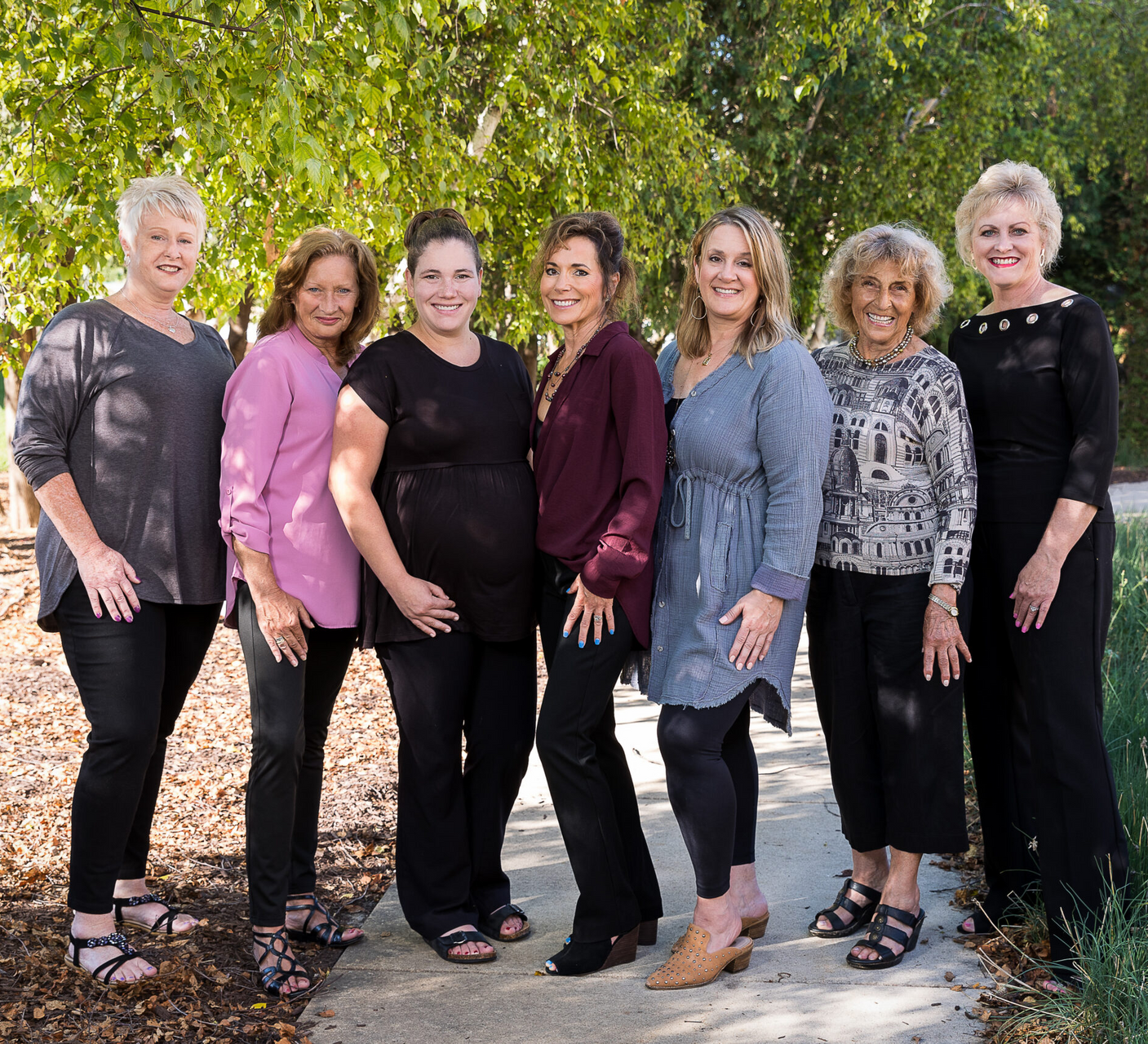 Seven people standing outdoors, smiling. They wear various colors of tops and black pants.