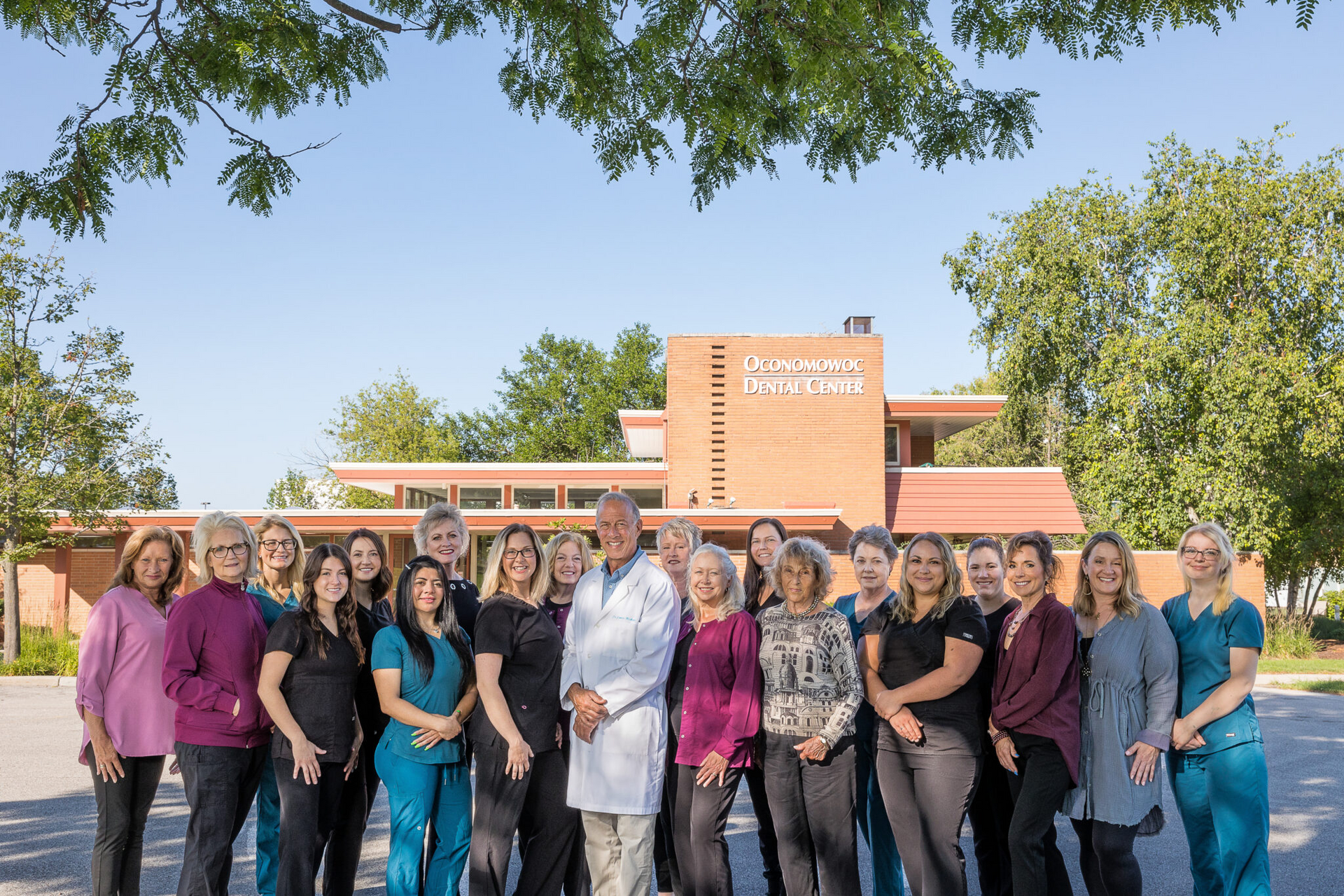 Group of people posing outside a brick building. Many are wearing scrubs; some are smiling. Sunny day.
