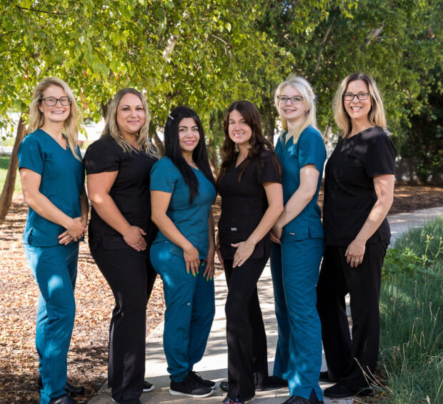 Six people in scrubs posing outdoors. Some in teal, some in black, near a tree.