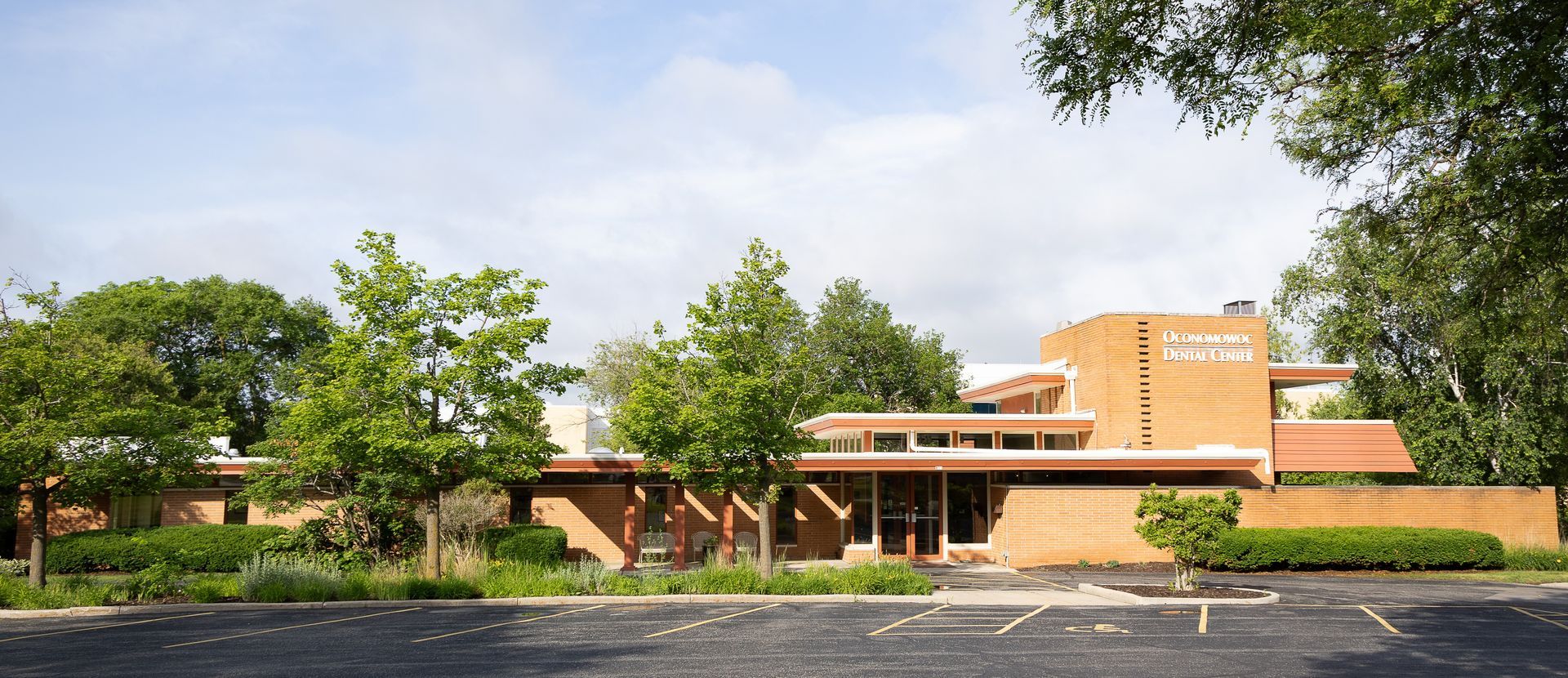 Brick building with a curved tower behind trees and a parking lot in front under a clear sky.