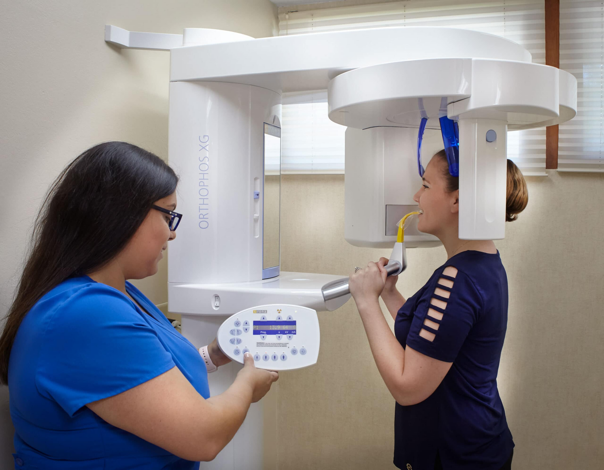Dentist using dental X-ray machine on patient; white machine, blue scrubs, dental office setting.