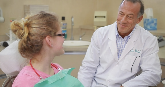 Dentist smiles at a seated patient in an examination room. Pink shirt, white lab coat.