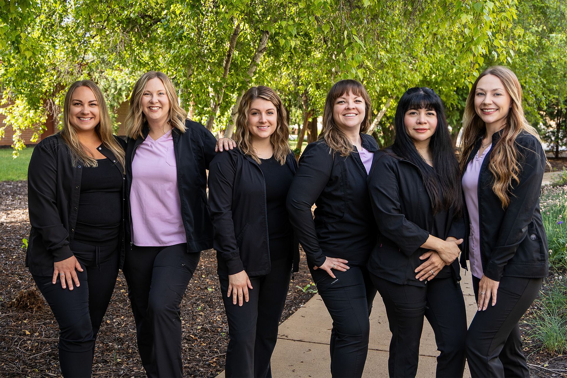 Six women in black outfits standing together outdoors in front of green trees.