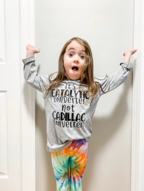 A little girl wearing a shirt that says `` catalyst converter not cadillac inverter '' is standing in front of a door.