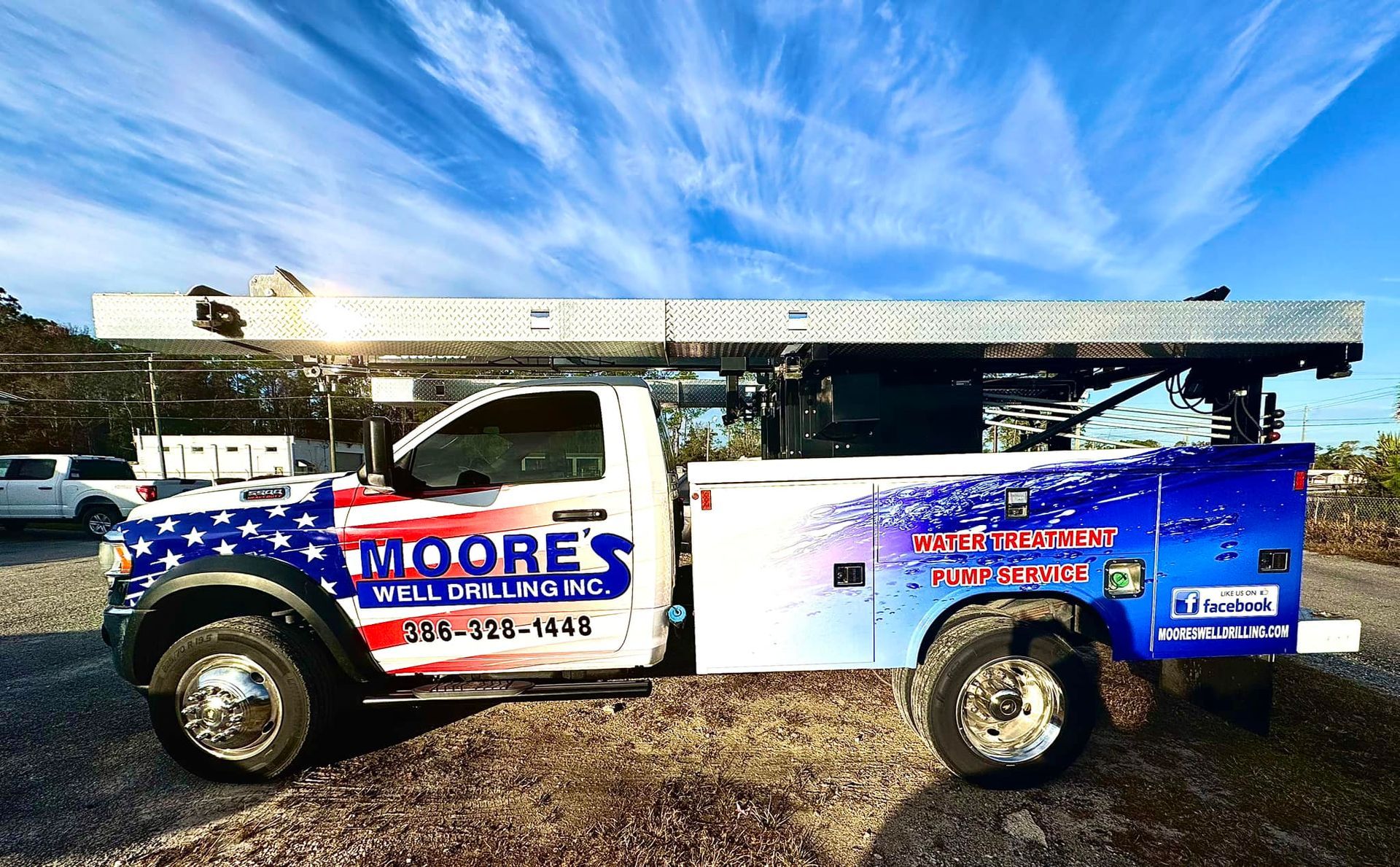 Moore's Well Drilling truck with American flag design on the side, parked on a gravel lot under a cloudy blue sky.