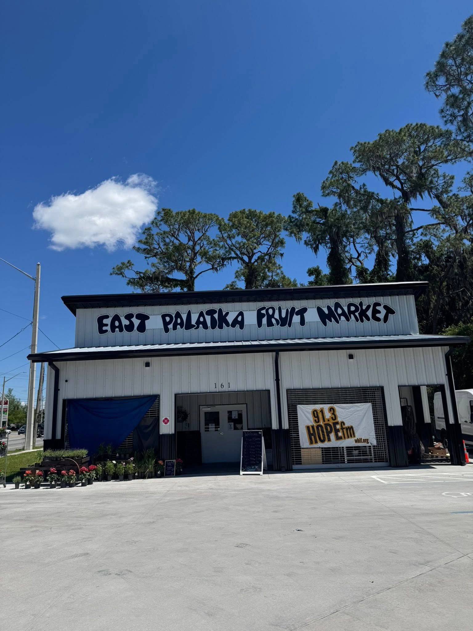 East Palatka Fruit Market, a white building with black lettering and a blue shutter, under a blue sky with clouds.