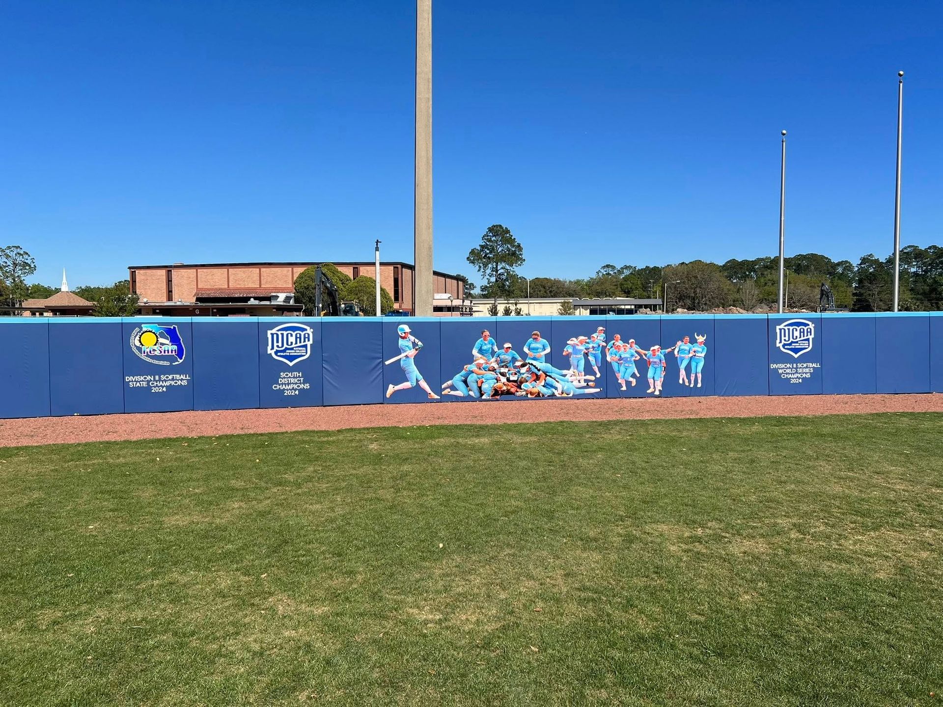 Blue baseball outfield wall with a team mural and logos; green grass in front and blue sky above.