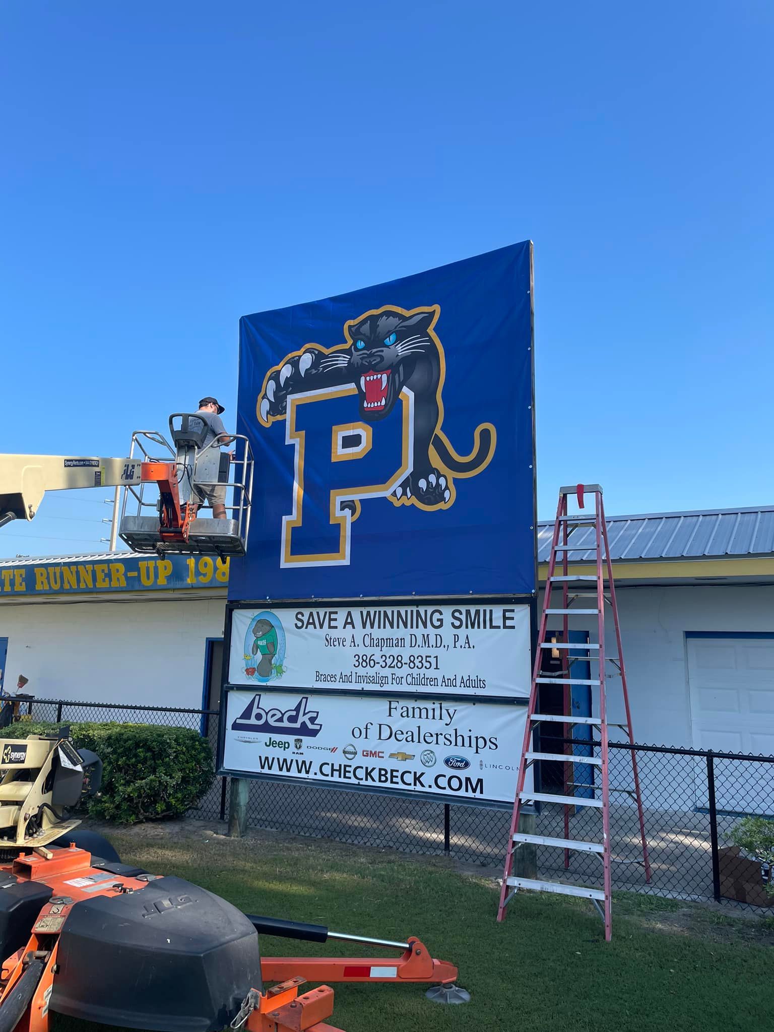 A worker on a lift installing a blue sign with a panther mascot and the letter 