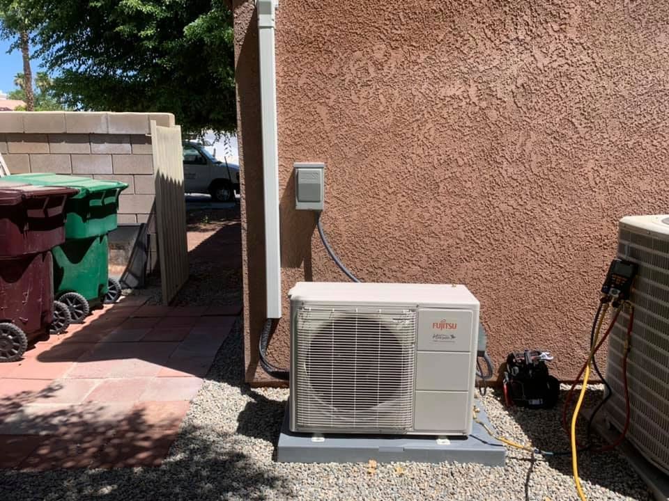 A white air conditioner is sitting outside of a building next to a trash can.