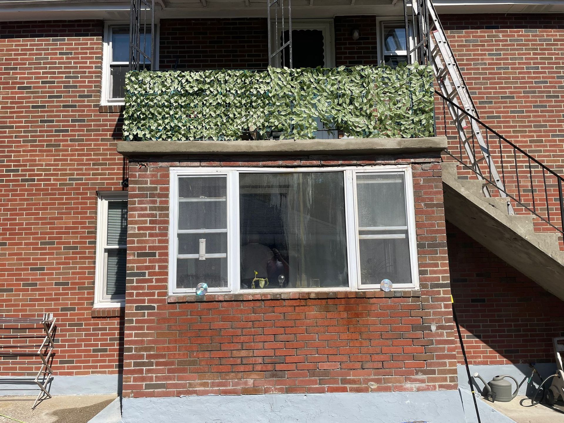Red brick building with a balcony covered in green plants, windows below. Staircase to the right.