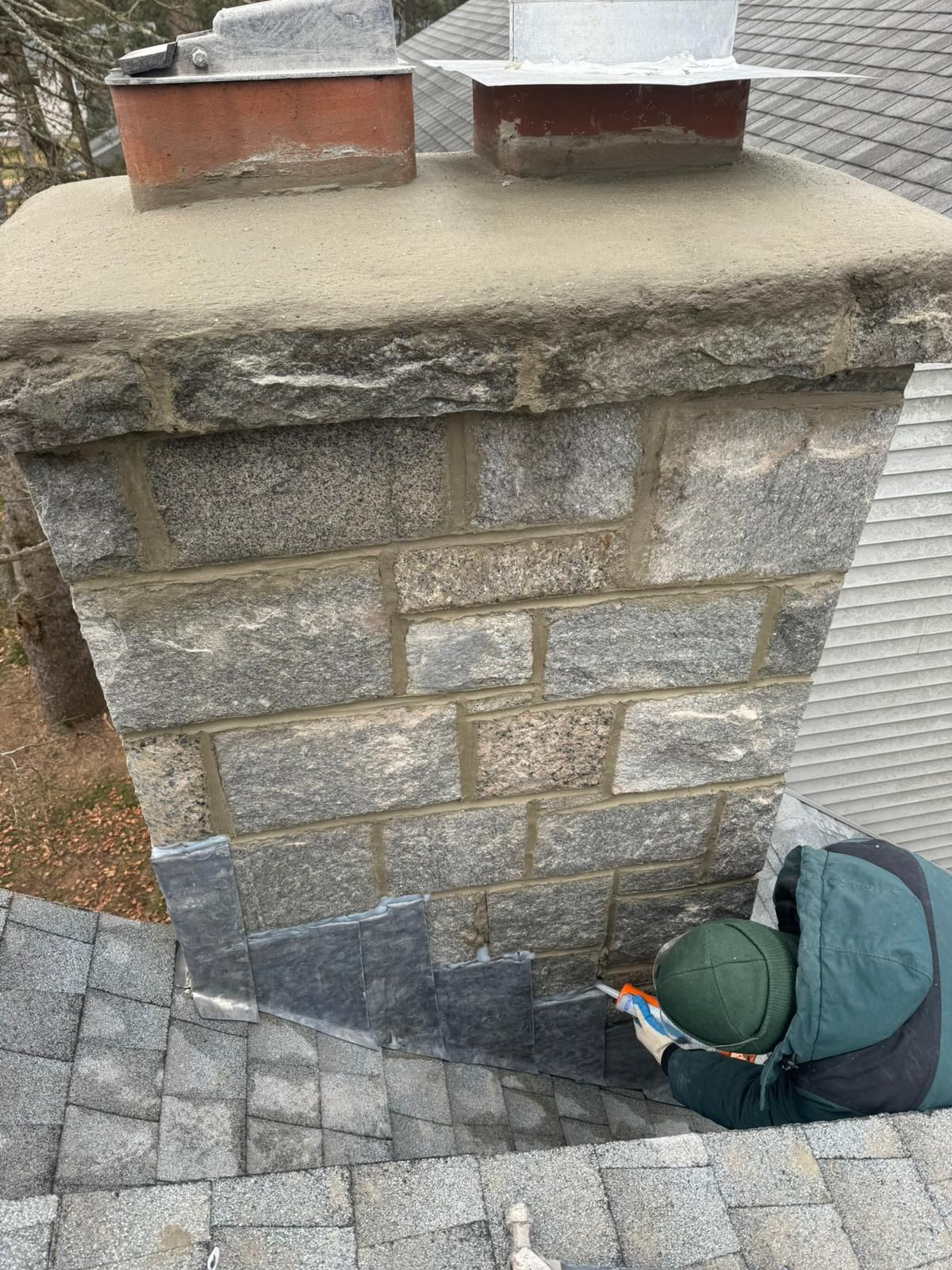 Person repairs chimney flashing on a gray shingled roof. Stone chimney has two flue tops.
