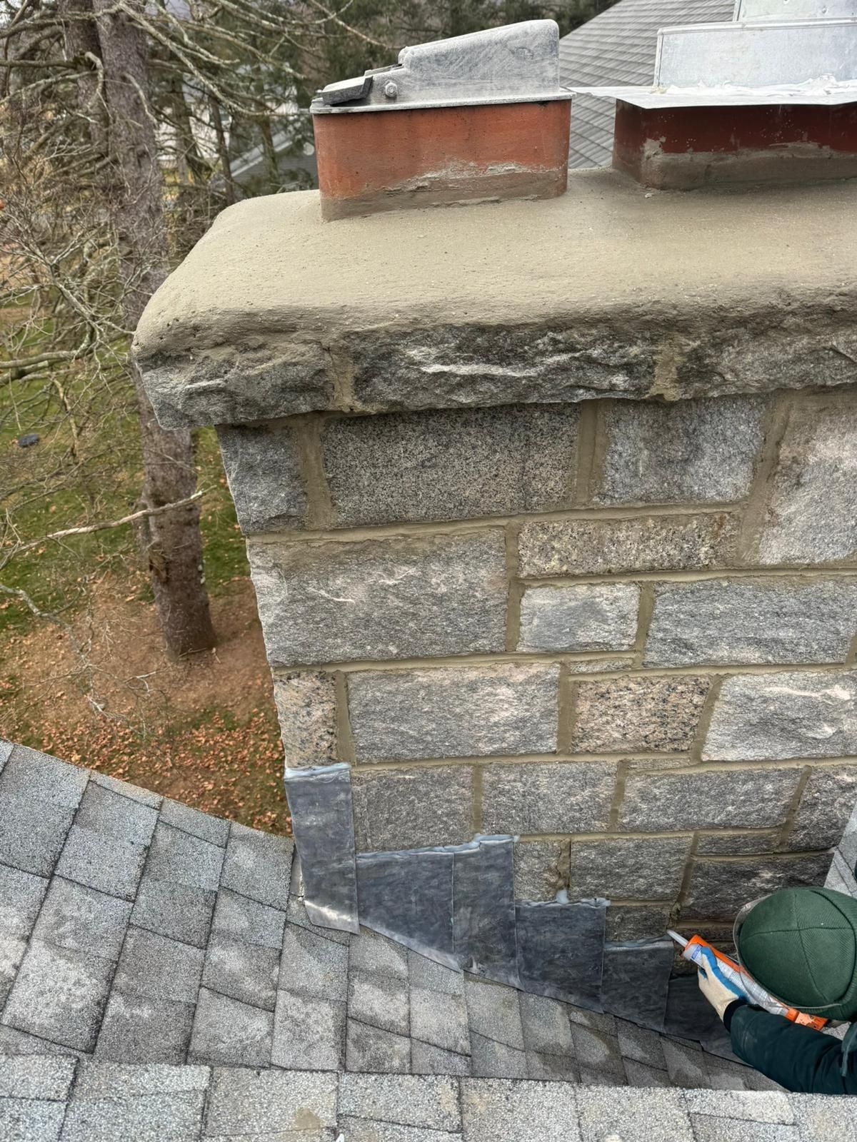 Chimney on a roof. Granite block construction, topped with concrete cap and brick flue. Shingles and a person visible.