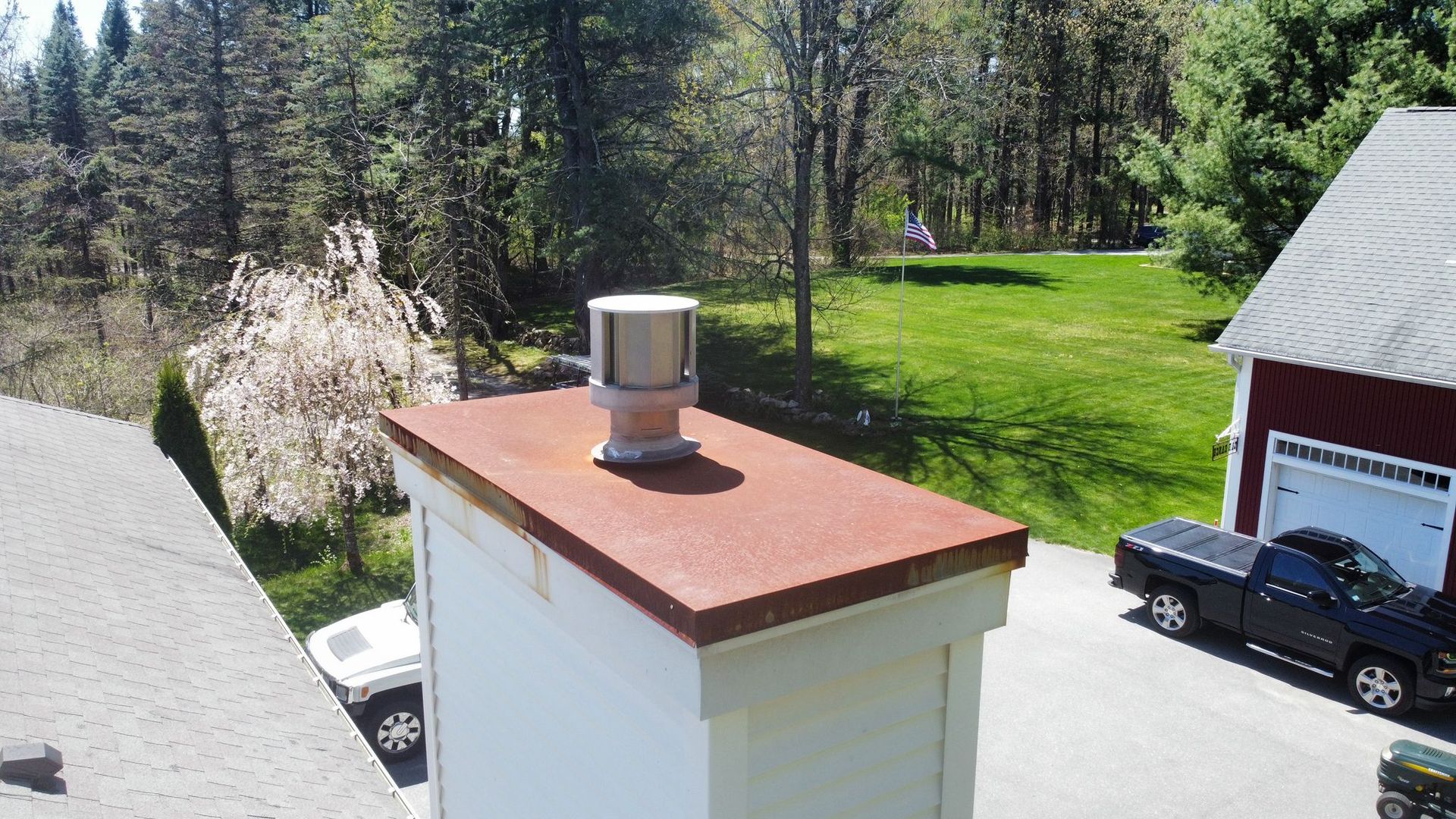 Chimney with a metal cap on a house roof, surrounded by trees and a lawn.