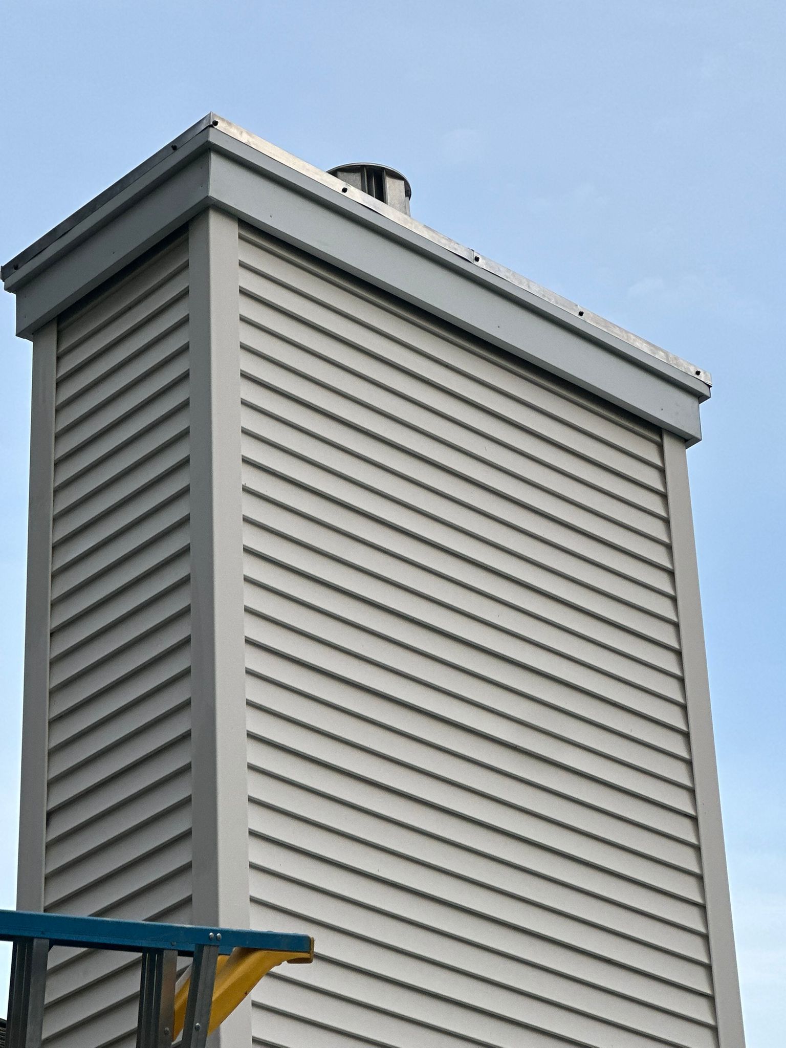 Tall, rectangular chimney with light gray siding and a dark chimney cap against a blue sky.