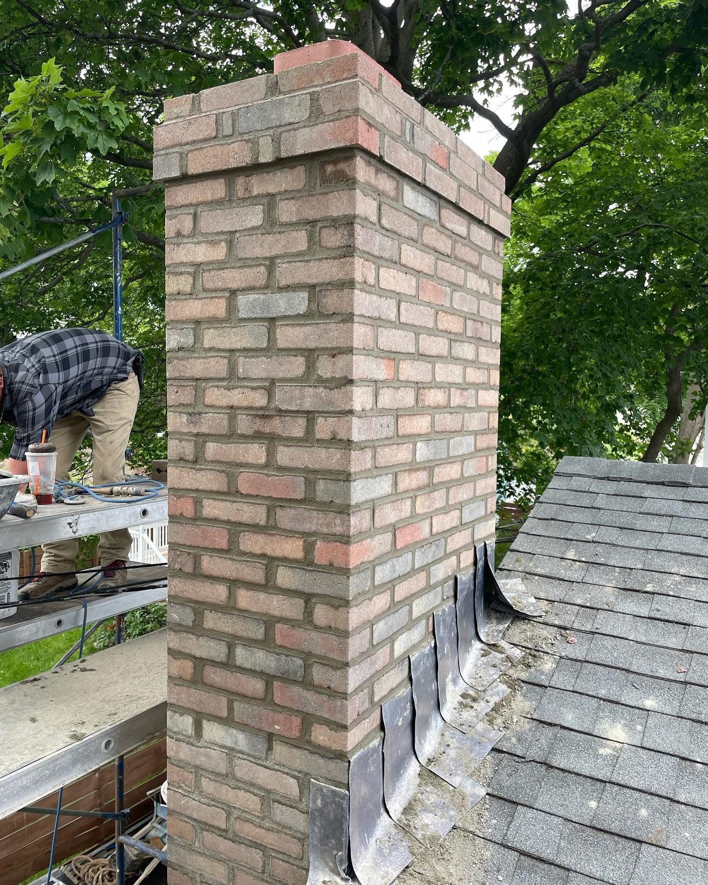 Brick chimney repair on a rooftop; a worker in a plaid shirt is visible on scaffolding.