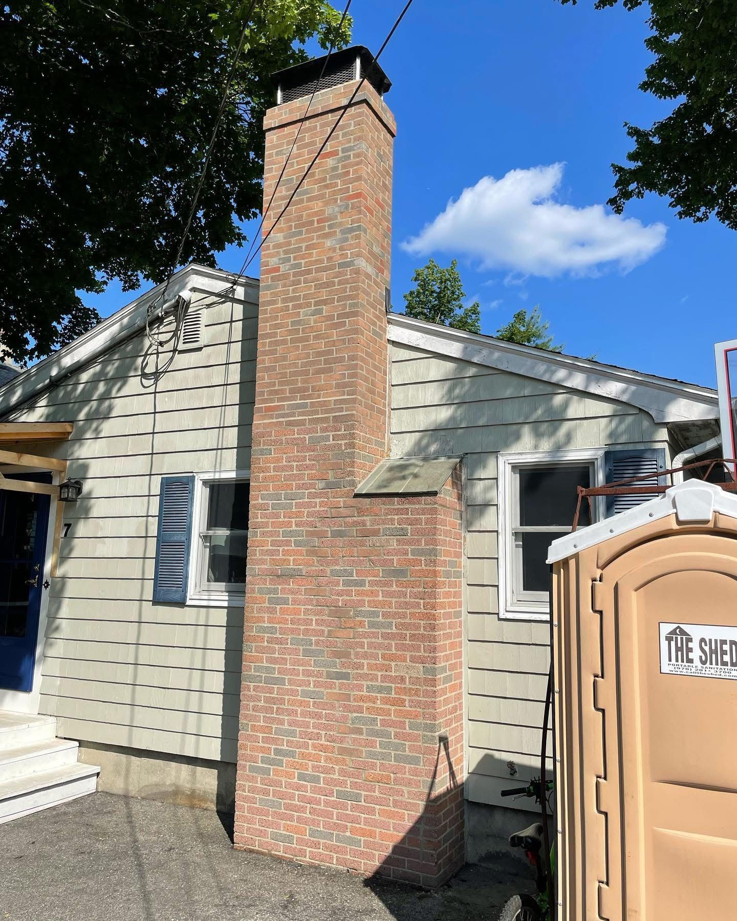 Tall brick chimney attached to a light-colored house; a portable toilet is next to the house.
