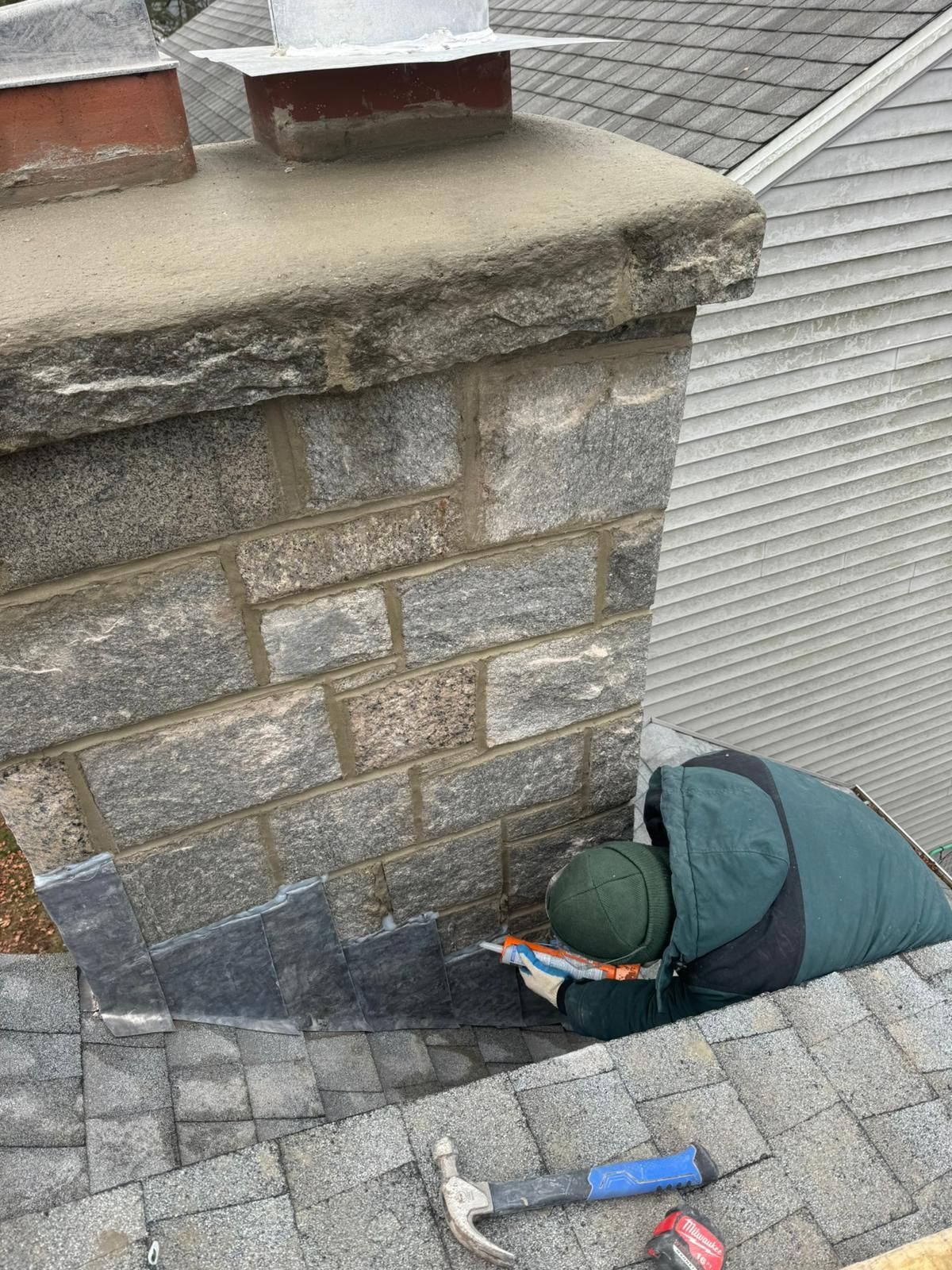 Person repairing roof flashing near a stone chimney.