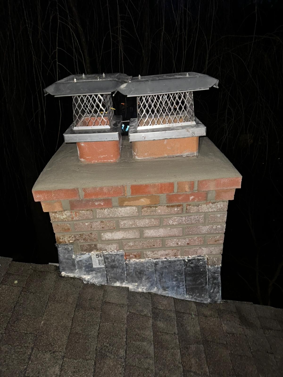 Brick chimney with two metal chimney caps on a dark roof at night.