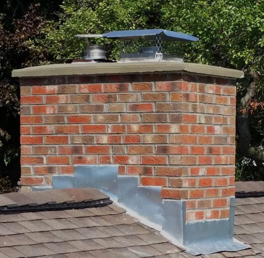 Brick chimney on a roof with metal flashing and cap. Green trees in the background.