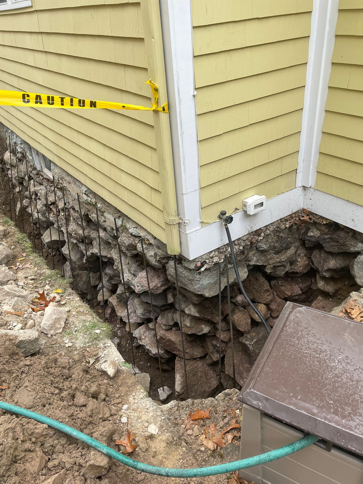 Exterior house corner with damaged stone foundation, yellow caution tape, and exposed wiring.