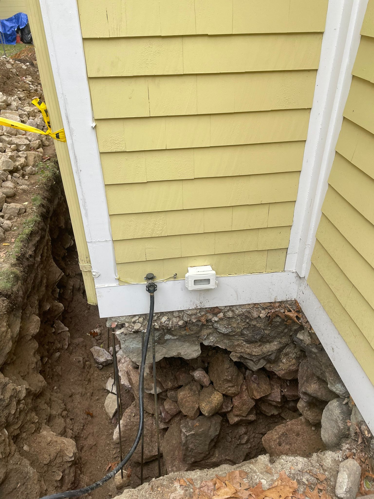 Trench dug next to a yellow building. Exposed foundation and electrical outlet visible.