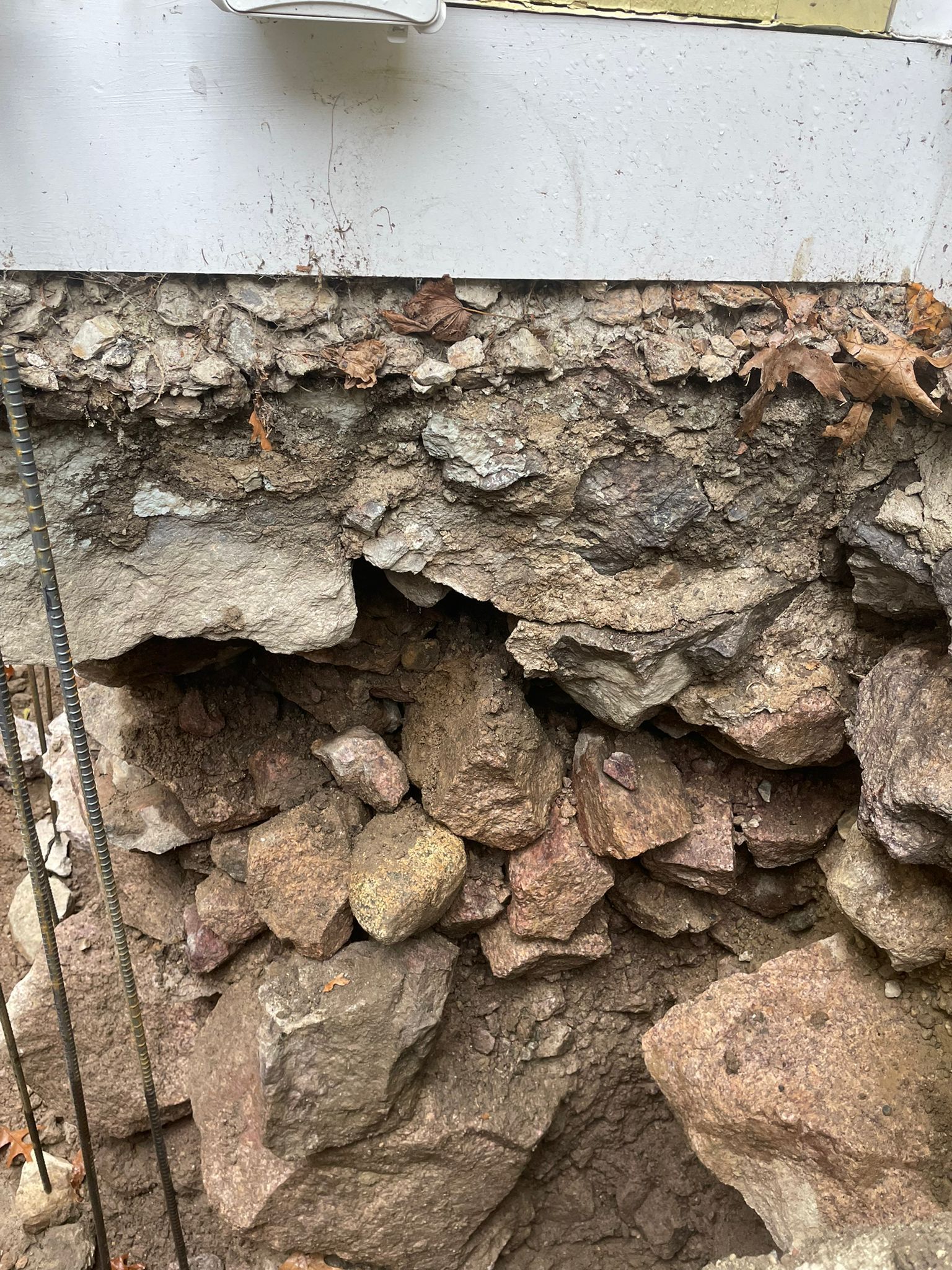 Damaged stone foundation of a building with exposed rocks, dirt, and cement under siding.