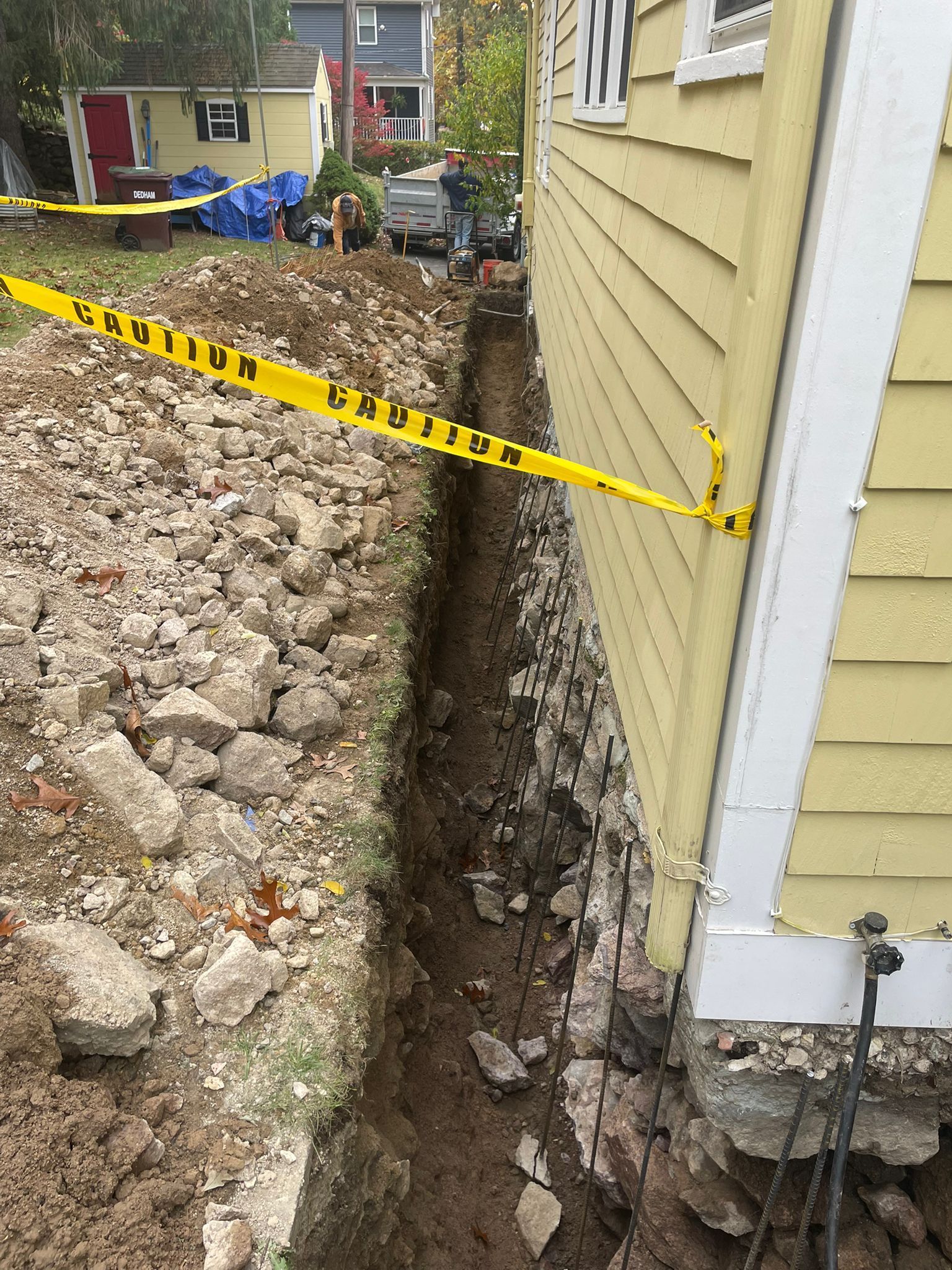 A trench dug next to a yellow house foundation, marked with caution tape, suggesting construction or repair work.
