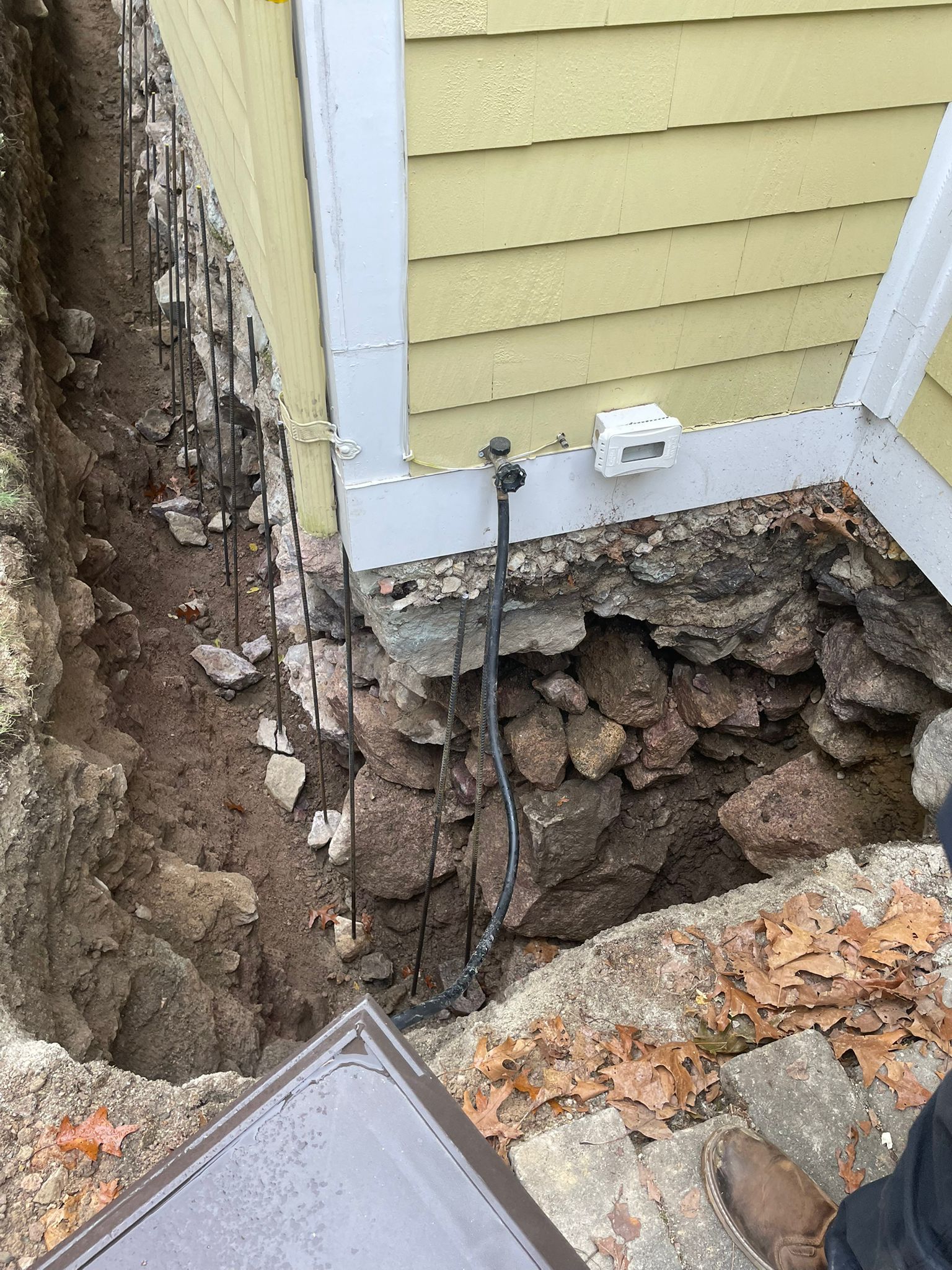 Trench dug along a building's foundation, exposing rocks and rebar, with a utility pipe visible.