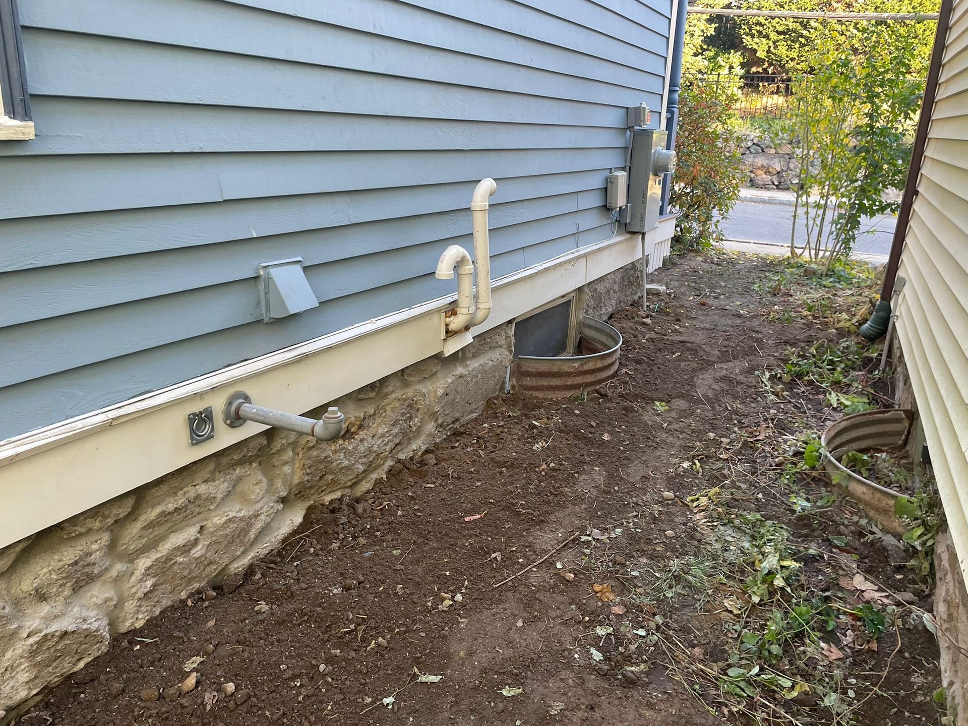 Side of a blue-sided house with a stone foundation. Brown dirt path runs along the wall. Metal pipes and electrical boxes are visible.