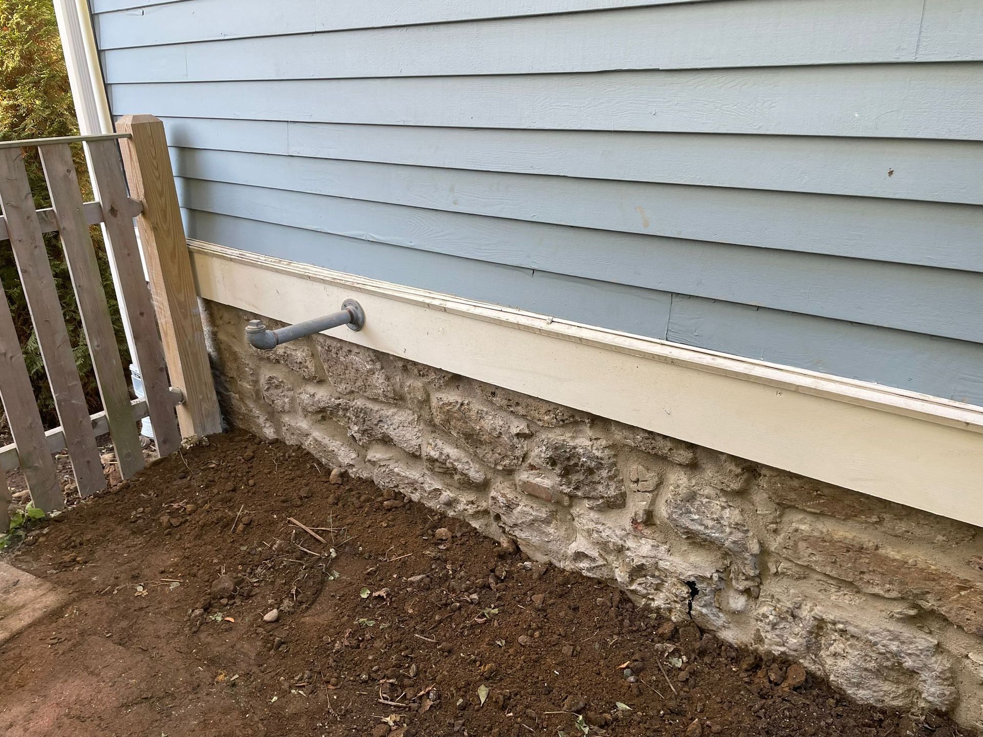 Stone foundation below blue siding, with a pipe and dirt. A fence is on the left.