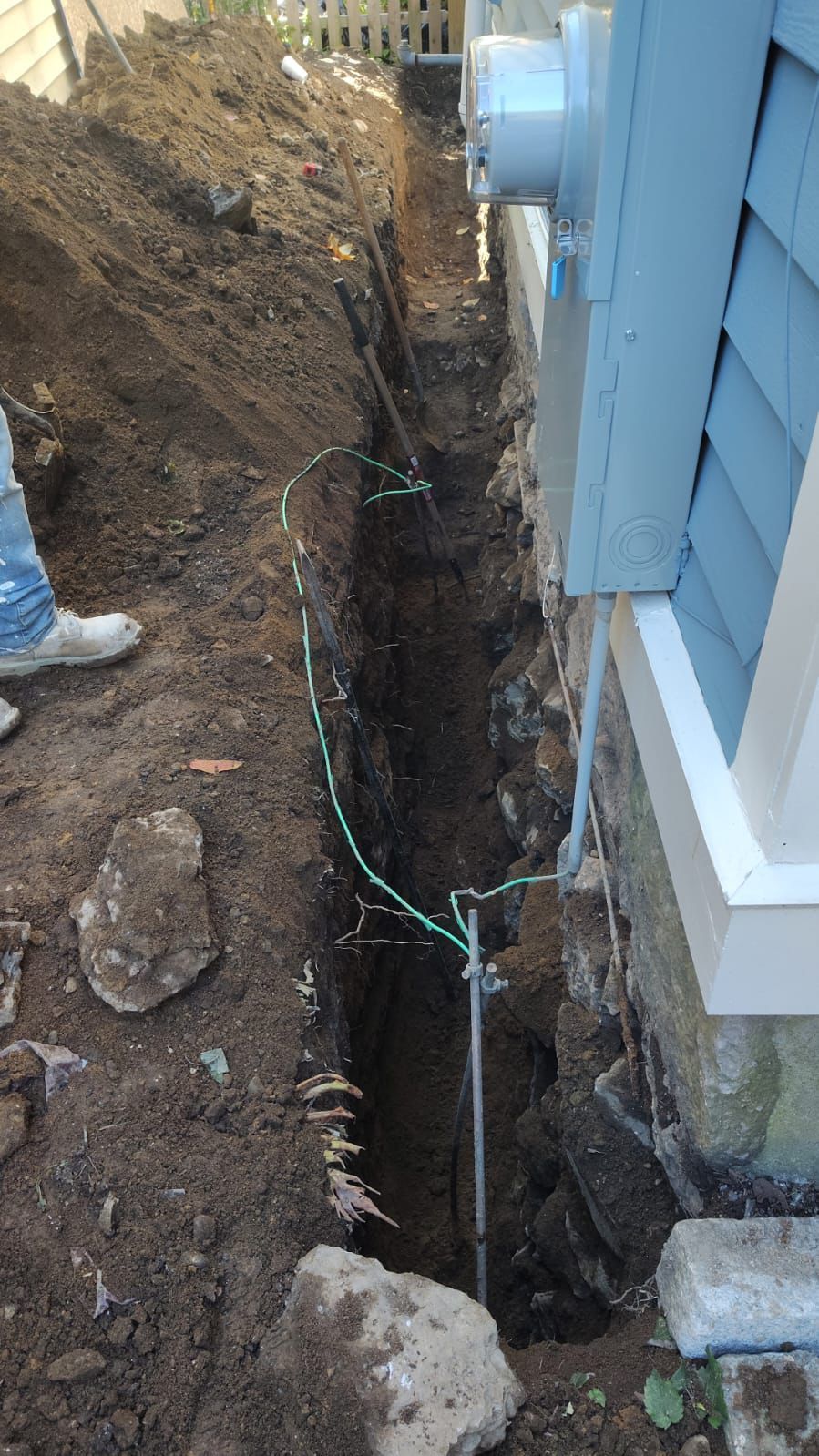 Trench dug next to a house foundation for electrical work. Green wire and metal grounding rod are visible.