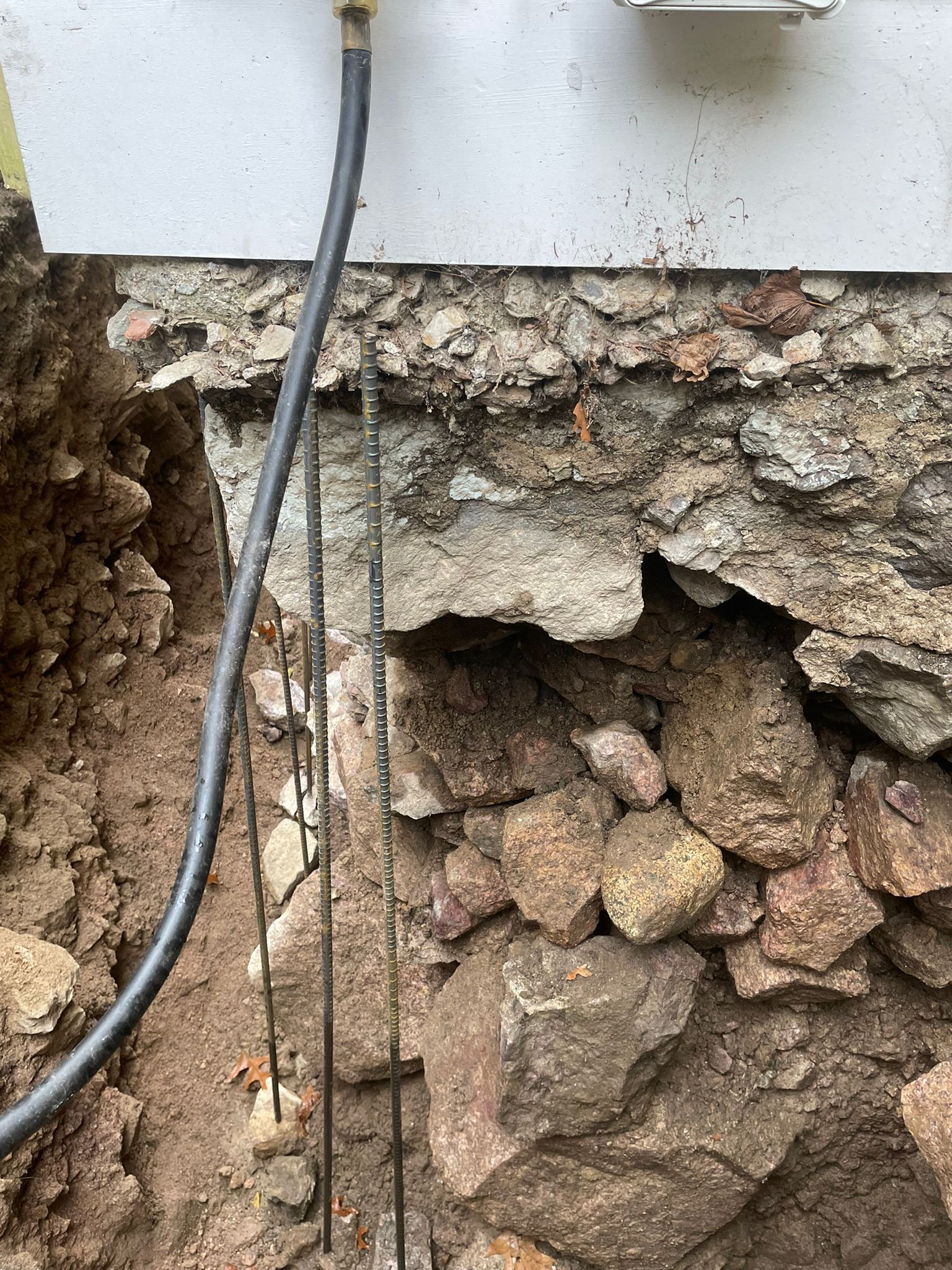Excavation revealing foundation made of stacked stones and concrete, with a black hose and metal rods visible.