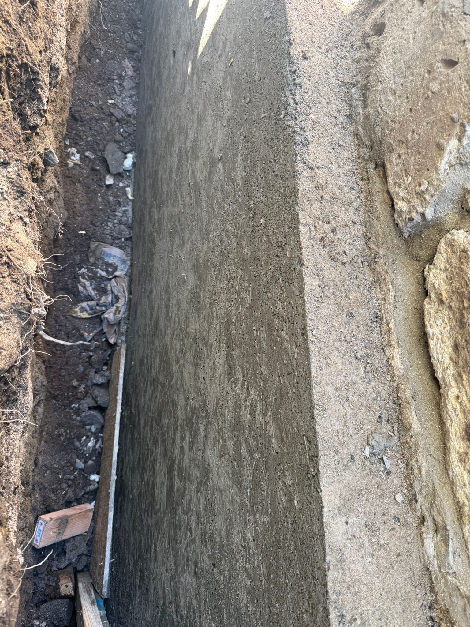 Trench with a concrete wall, dark soil, and sunlight.