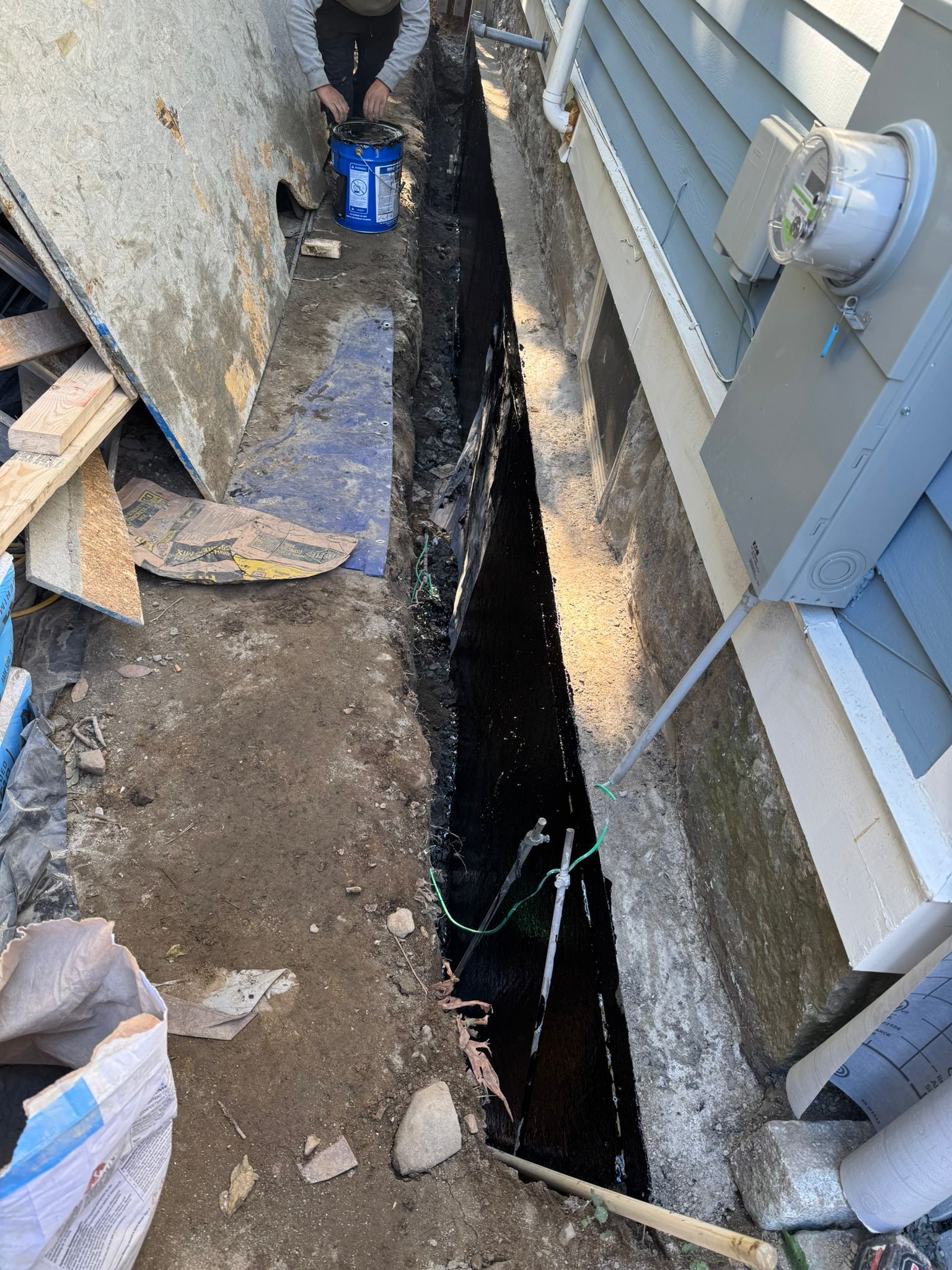 Narrow trench dug along a building's foundation. Person in the background. Construction debris and water present.