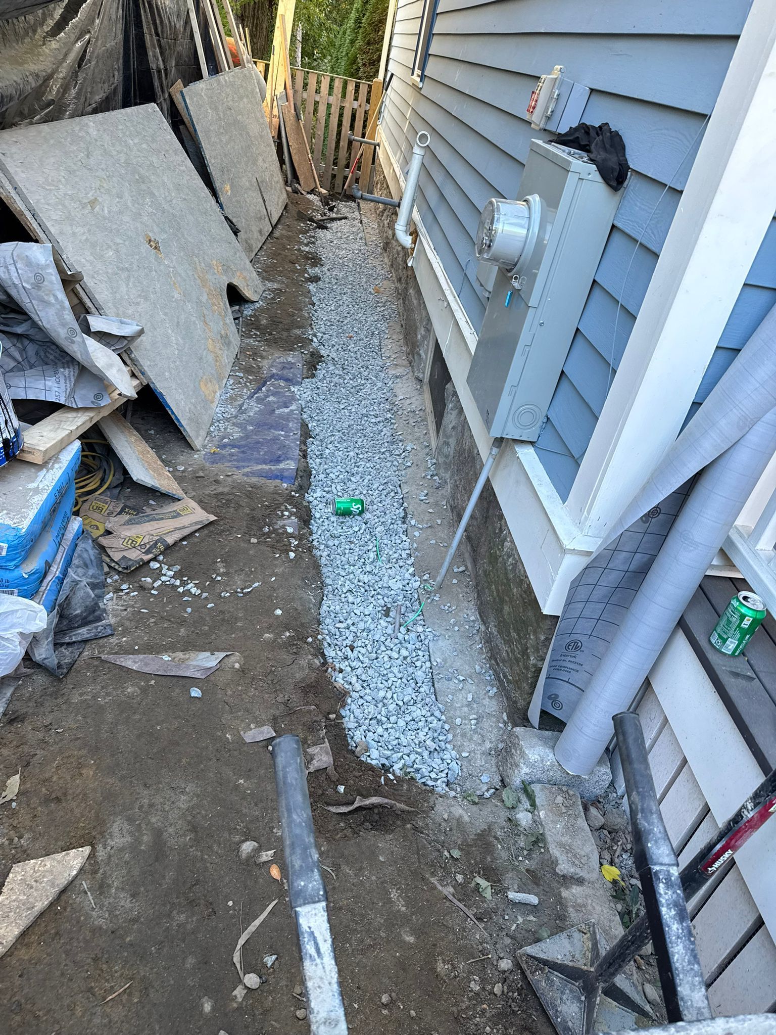Construction site next to a house with gravel drainage, grey siding, and debris.