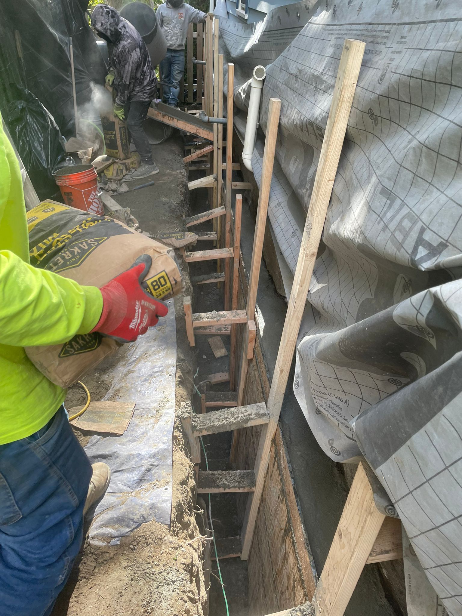 Construction worker pouring cement into a narrow trench with wooden supports.