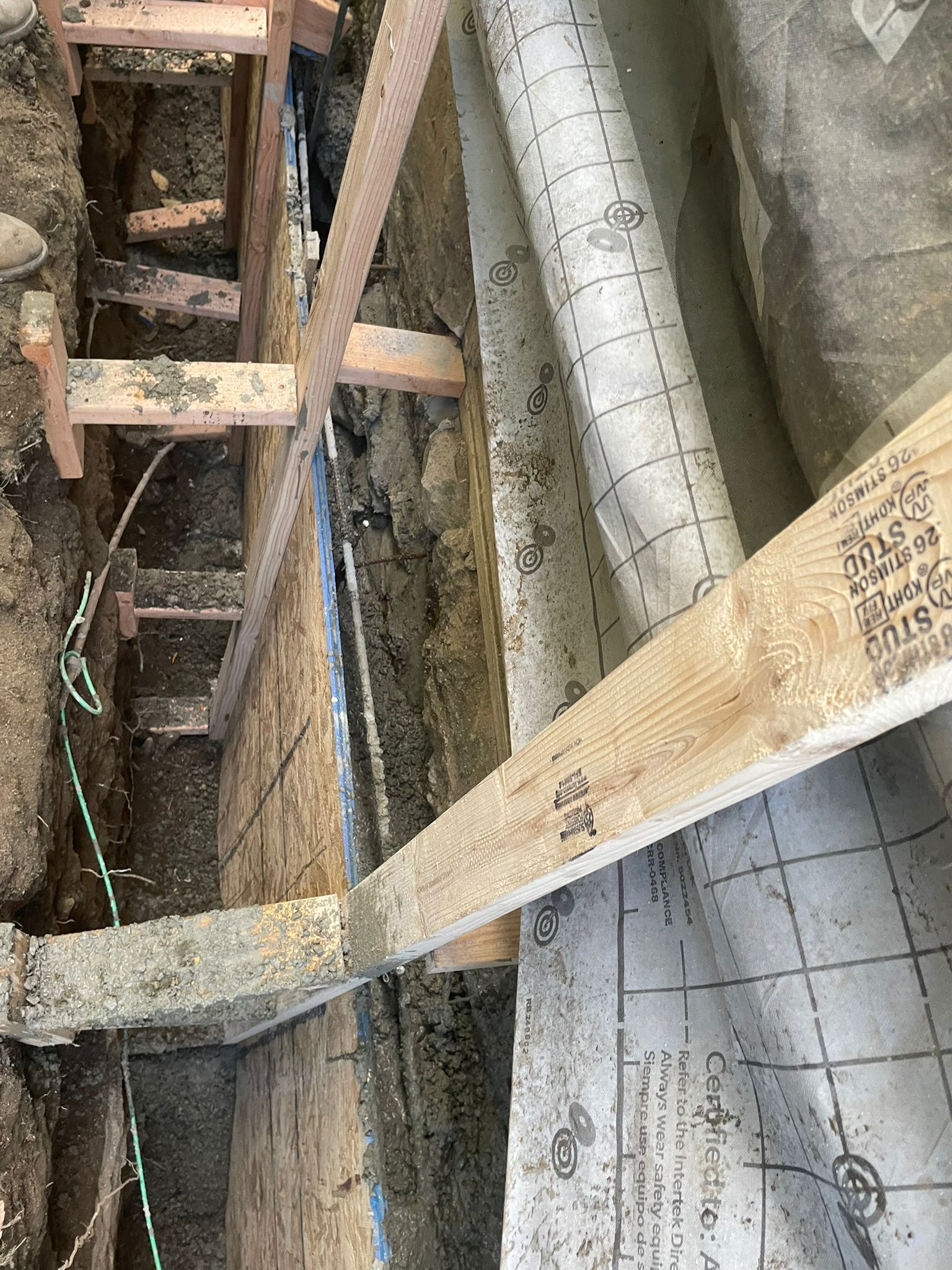 Construction site trench with wooden supports, plastic sheeting, and exposed earth.