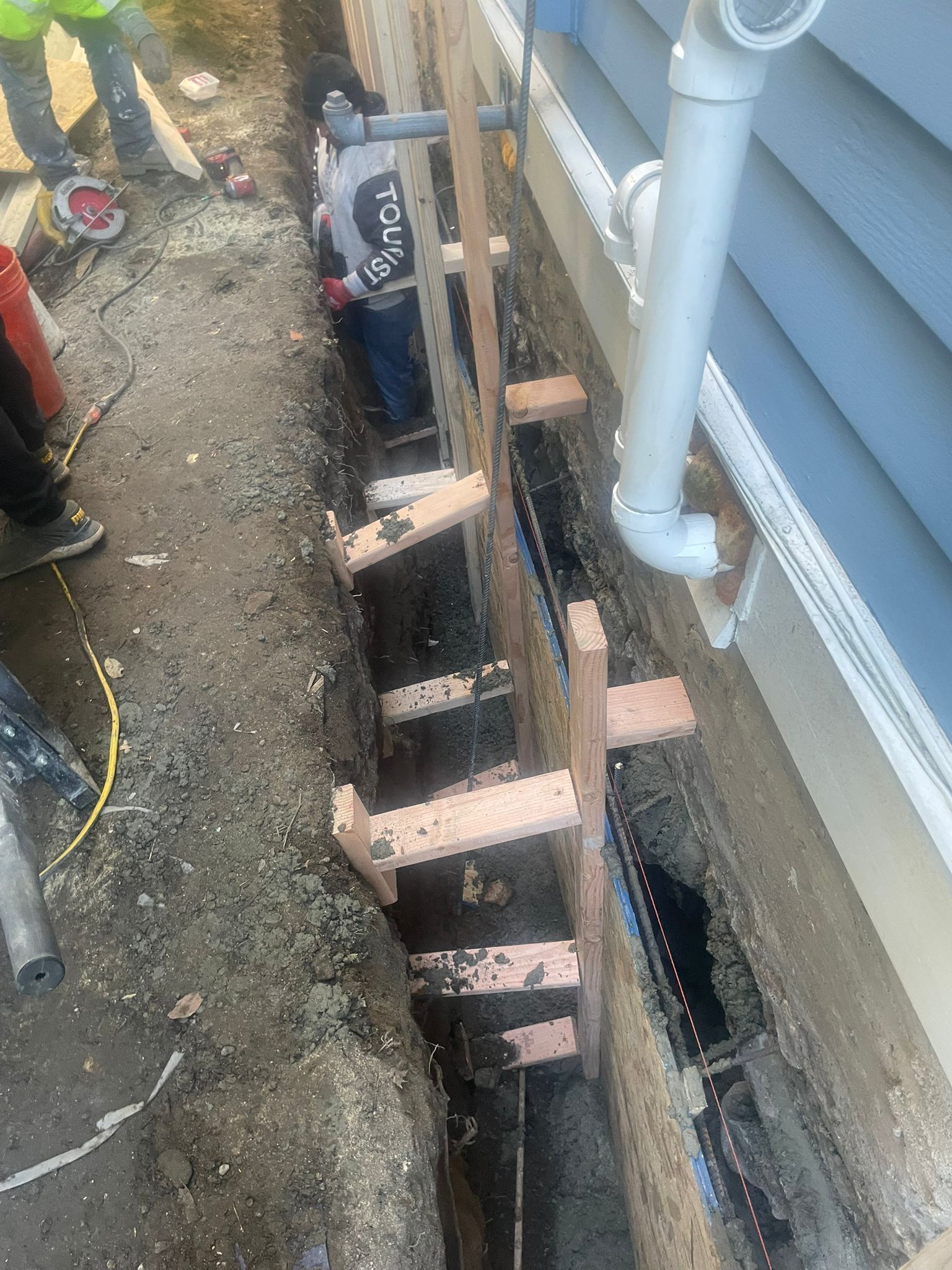 Construction workers in a trench next to a building, working on plumbing. Wood supports are in the trench.