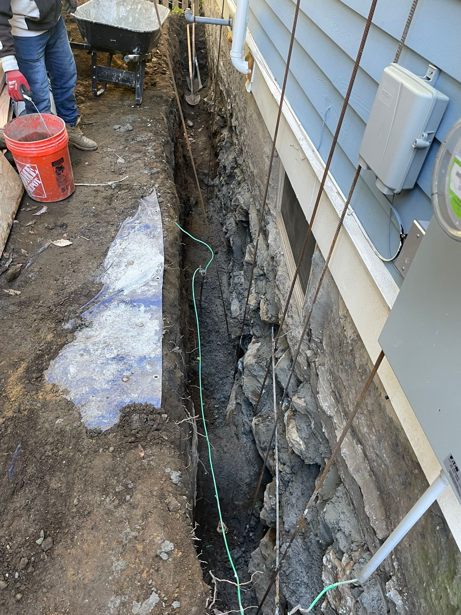 Trench dug along a light blue house wall with electrical wiring. A person and equipment visible.