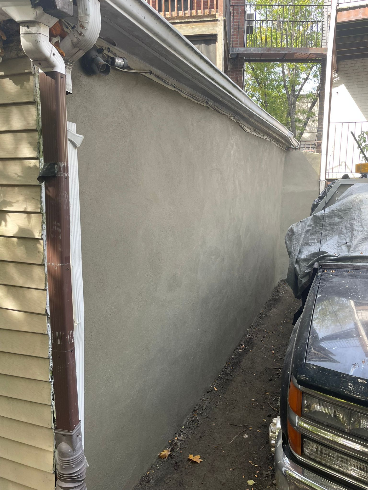 Gray stucco wall alongside a parked truck and a building with a downspout.