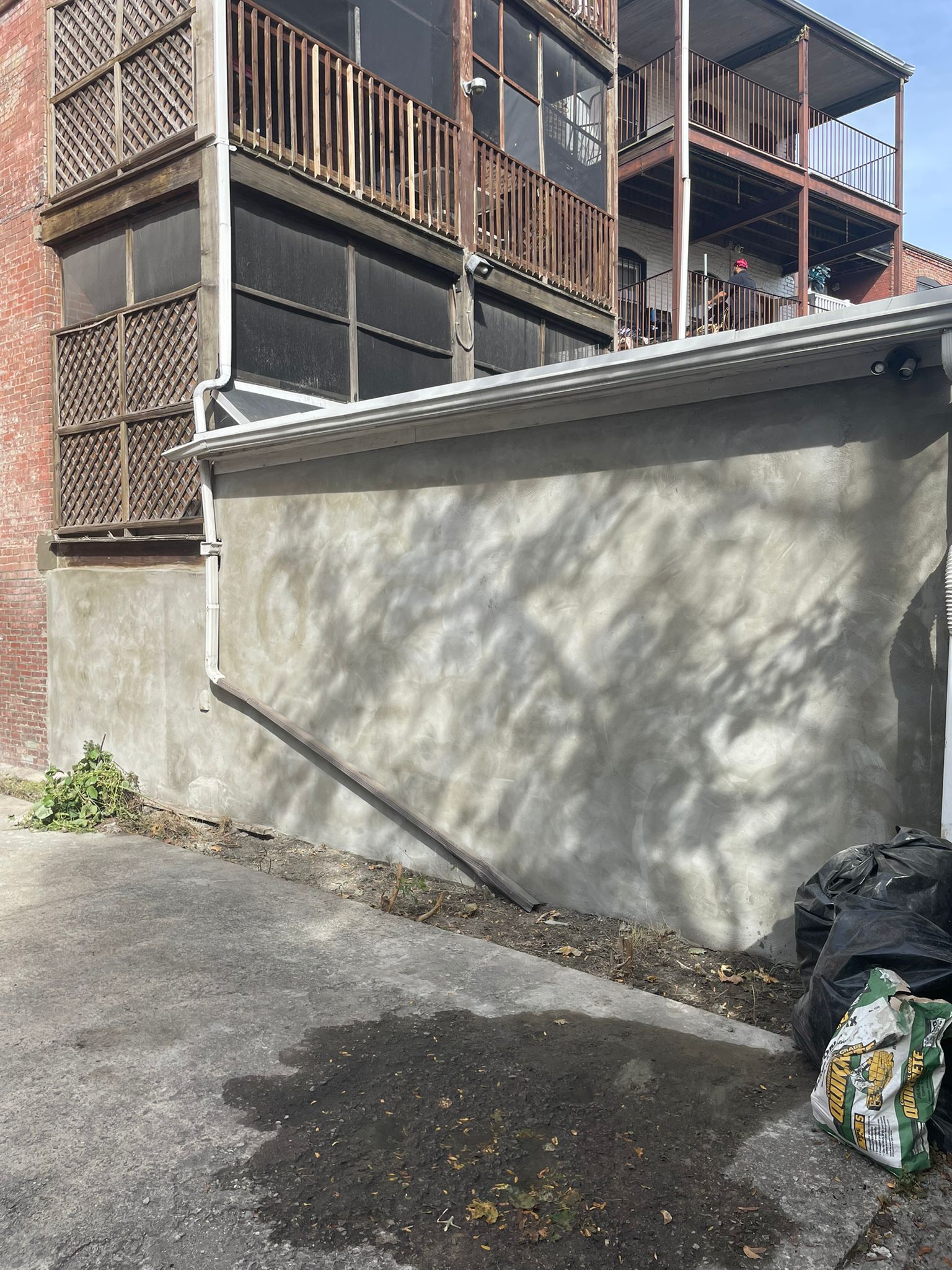 Back alley with concrete wall, building with balconies overhead, trash bags on ground.