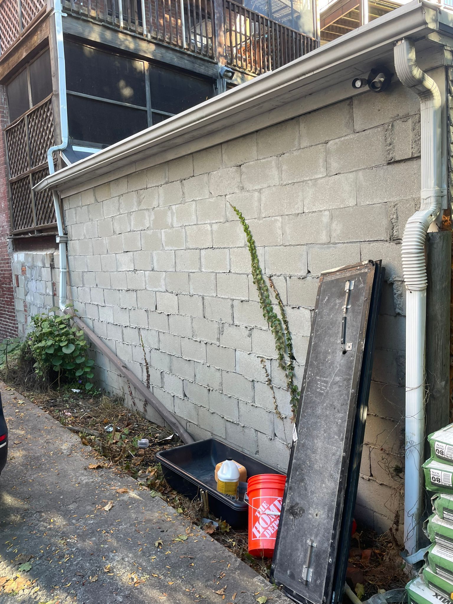 Concrete block wall with debris, gutter, and a climbing plant in an alley.