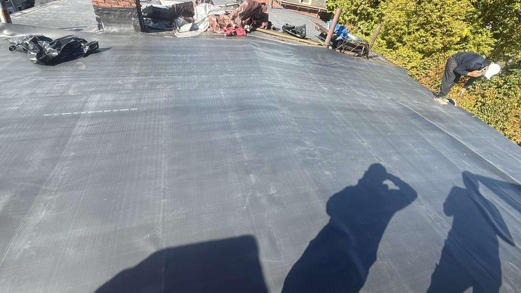 Person working on a flat, black roof on a sunny day. Shadow of the photographer is visible.