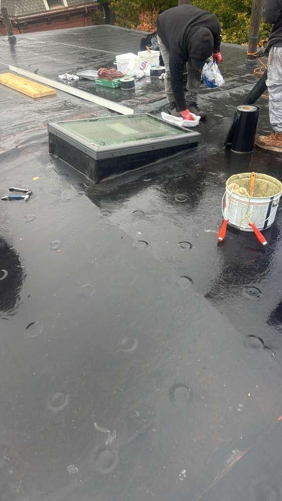 Workers on a black, wet roof. One near a skylight, the other holding a tool. Rain is visible.
