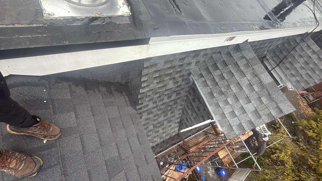 View of a roof with asphalt shingles, a metal flashing, and boots standing near the edge.