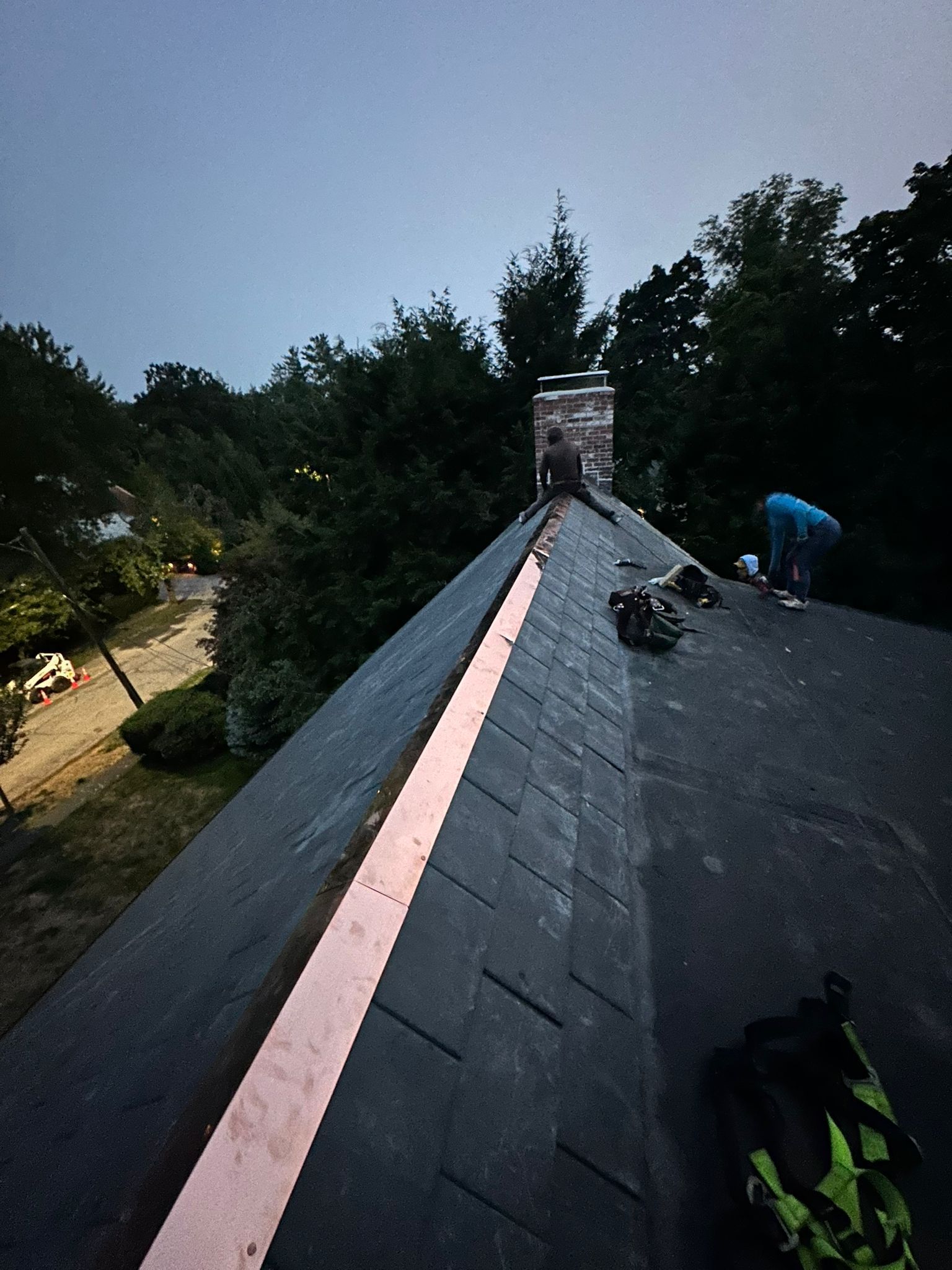 Workers on a dark roof with copper flashing. Trees and a chimney are in the background.