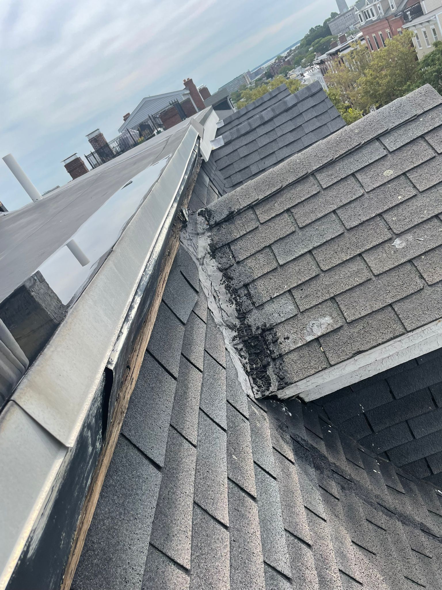Rooftop view of asphalt shingles, flashing, and flat roof with cityscape background under overcast sky.