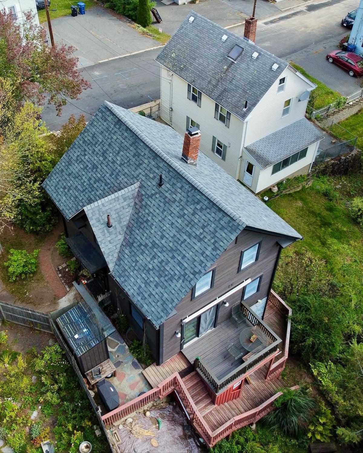 Aerial view of a brown house with a deck and a gray-roofed house in the background on a green, suburban lot.