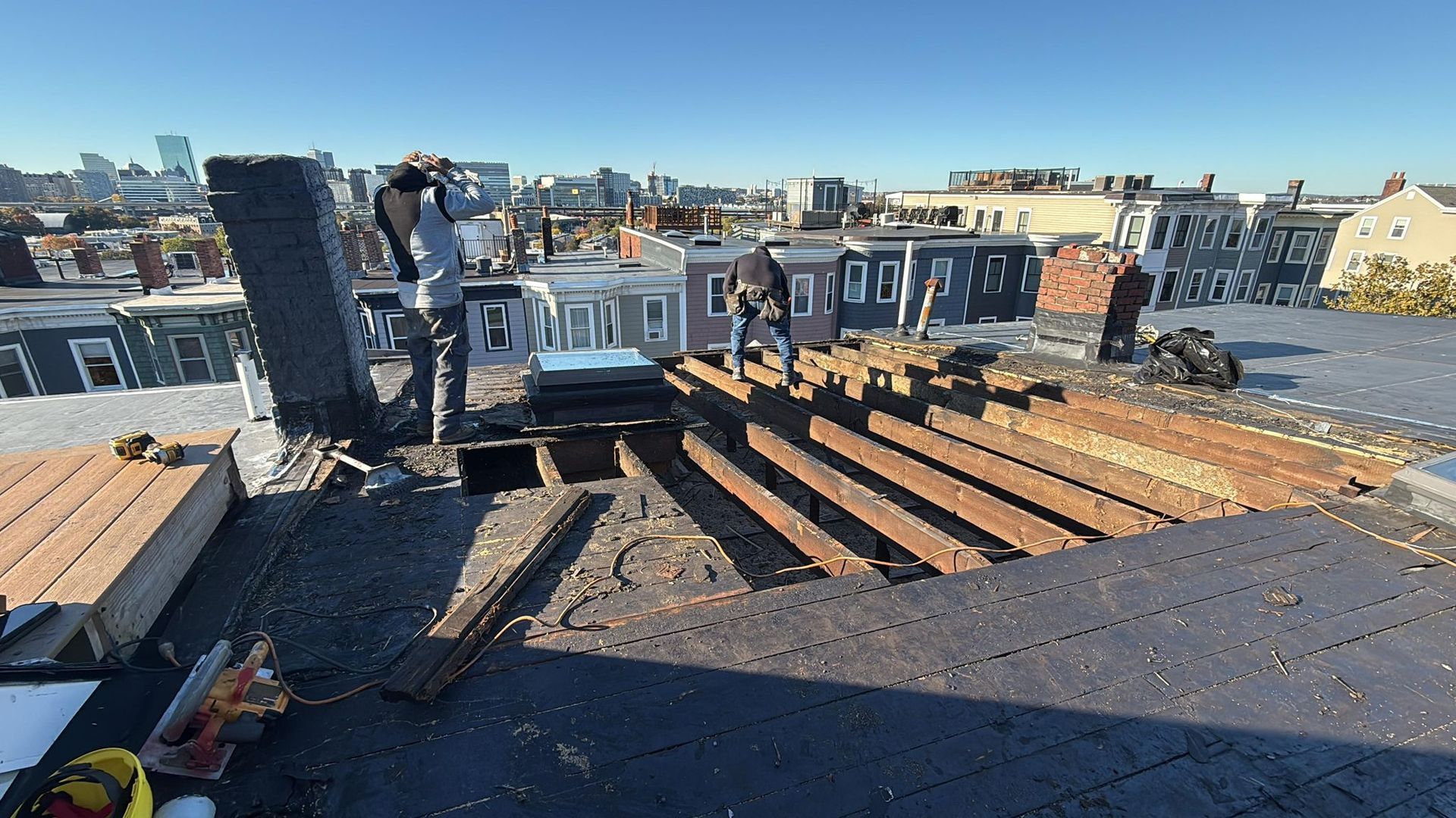 Workers on a city rooftop with exposed beams, dismantling part of the structure under a blue sky.